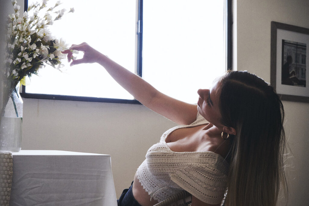 Marintia Bustos a model with long brown hair is captured in a serene moment by a window. She's reaching out to touch white flowers in a vase wearing a white lace top and the room has a minimalist elegant ambiance.